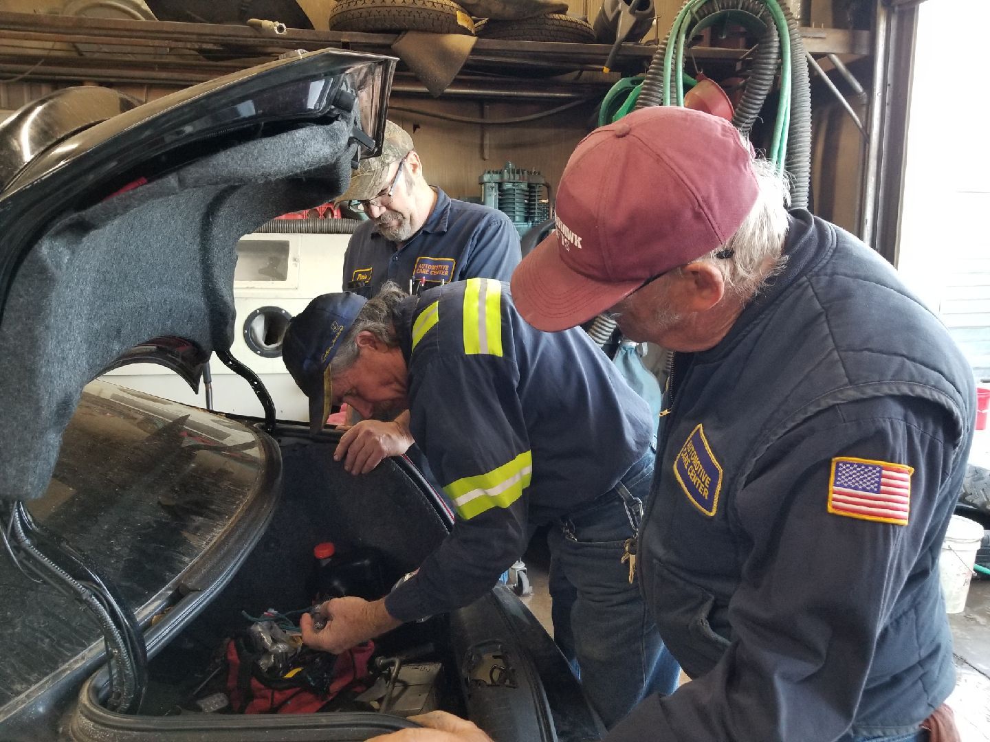 Three mechanics in uniforms examine the open trunk of a car inside a repair shop.