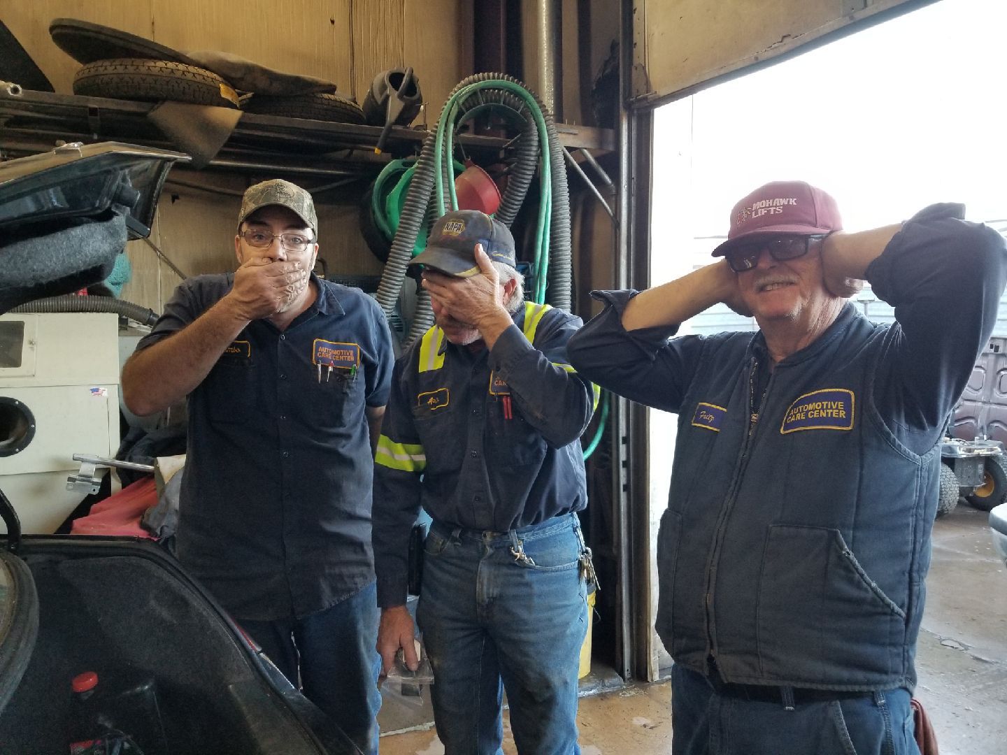 Three people in work uniforms standing in a garage, posing with hands over their mouths, eyes, and ears.