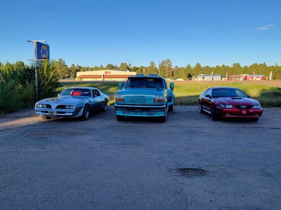 Three cars parked side-by-side in an outdoor lot on a sunny day: a silver Pontiac, a teal pickup truck, and a red coupe.