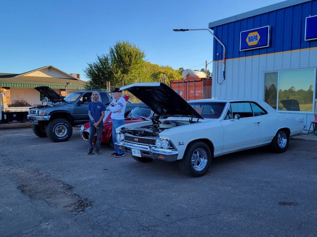 Two people stand between a dark truck and a white classic car with open hoods in front of an NAPA Auto Parts store.