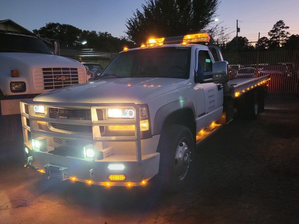 A white flatbed tow truck with glowing amber warning lights and a brush guard parked in a lot at dusk.