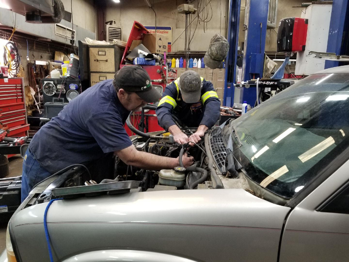 Two mechanics in a shop work together under the open hood of a silver car.