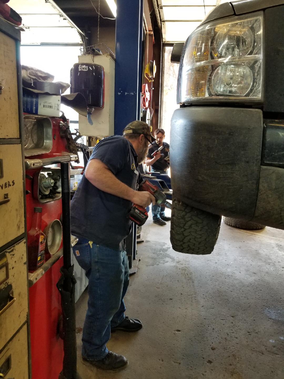 A mechanic using a power tool to work on a vehicle elevated on a lift inside an auto repair shop.
