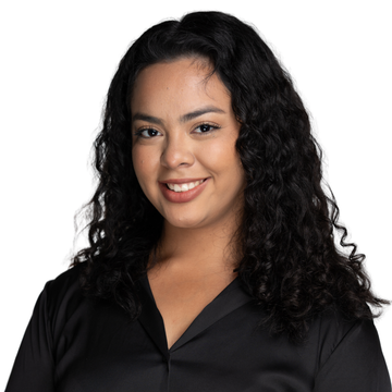 Woman with curly black hair smiles, leaning against a tree in a park; wearing a white top.