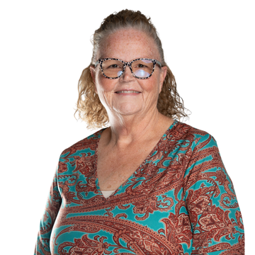 Woman with glasses, wearing a pink lace top, smiles at camera, outdoors with tree in background.