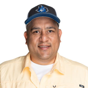 Man with a shaved head, smiling, outdoors near a tree, wearing a button-down shirt.