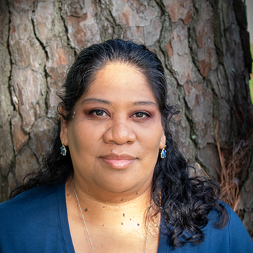 Woman with dark hair in blue top, in front of tree trunk, looking at the camera.