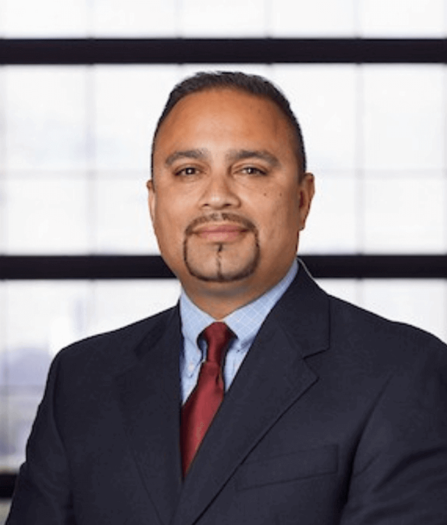 Man in a suit with a goatee, red tie, against a window background.