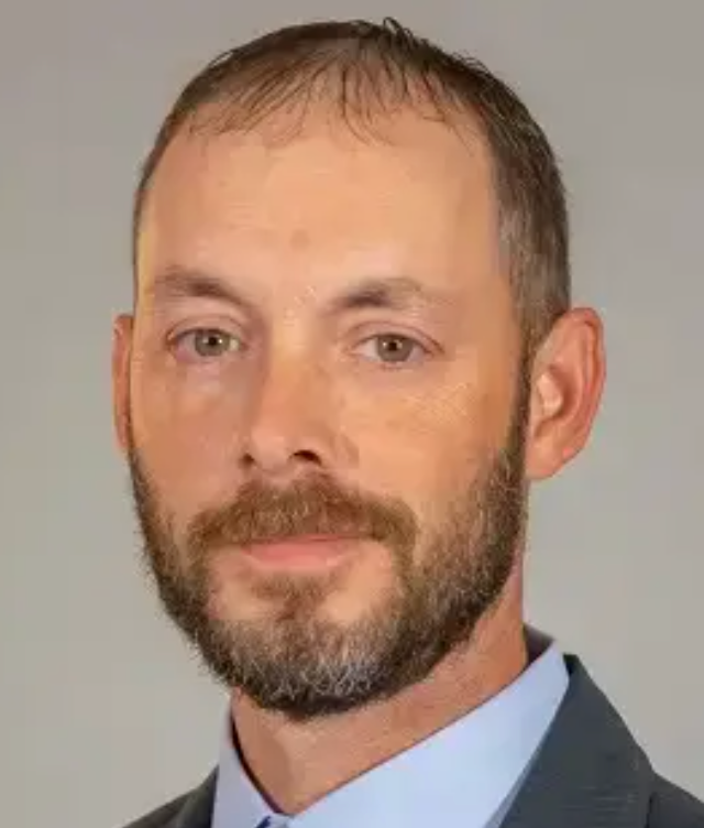 A middle-aged man with short brown hair and beard, wearing a blue shirt and blazer, looking at the camera.