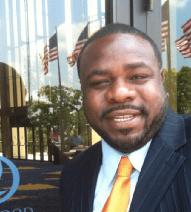 Man in suit smiling in front of a building with American flags.