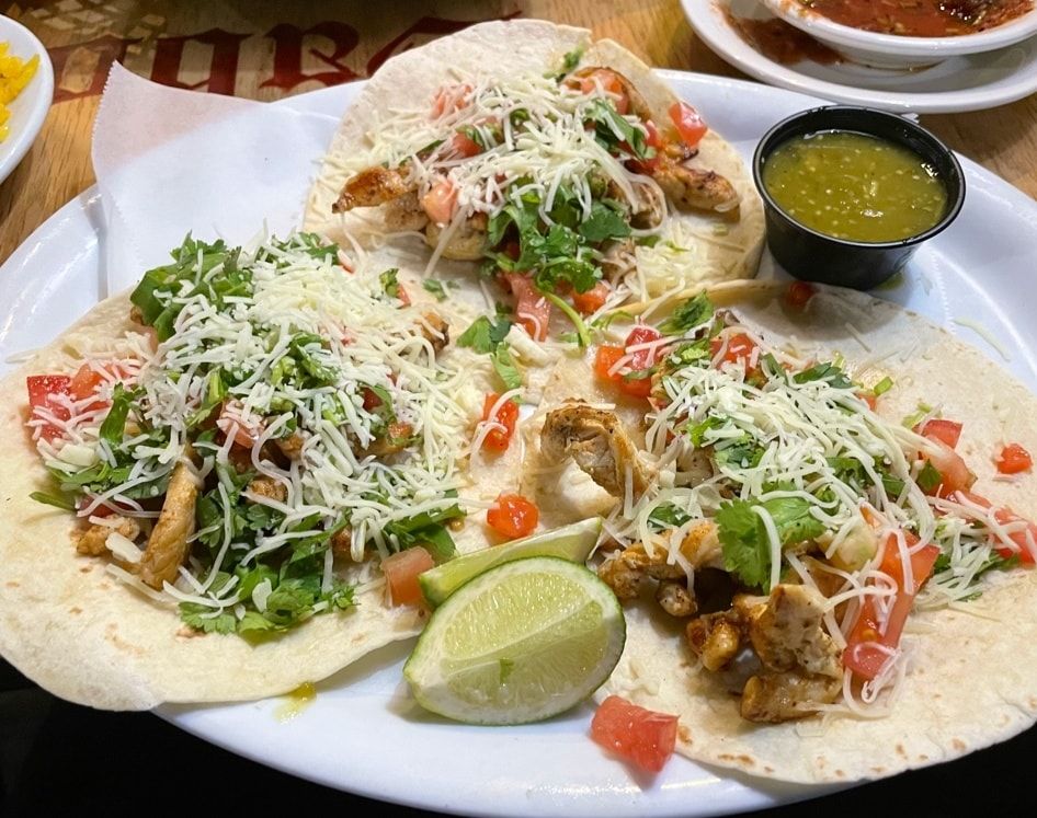 Plate of tacos topped with cheese, lettuce, tomatoes, and lime wedges, with green salsa on the side.