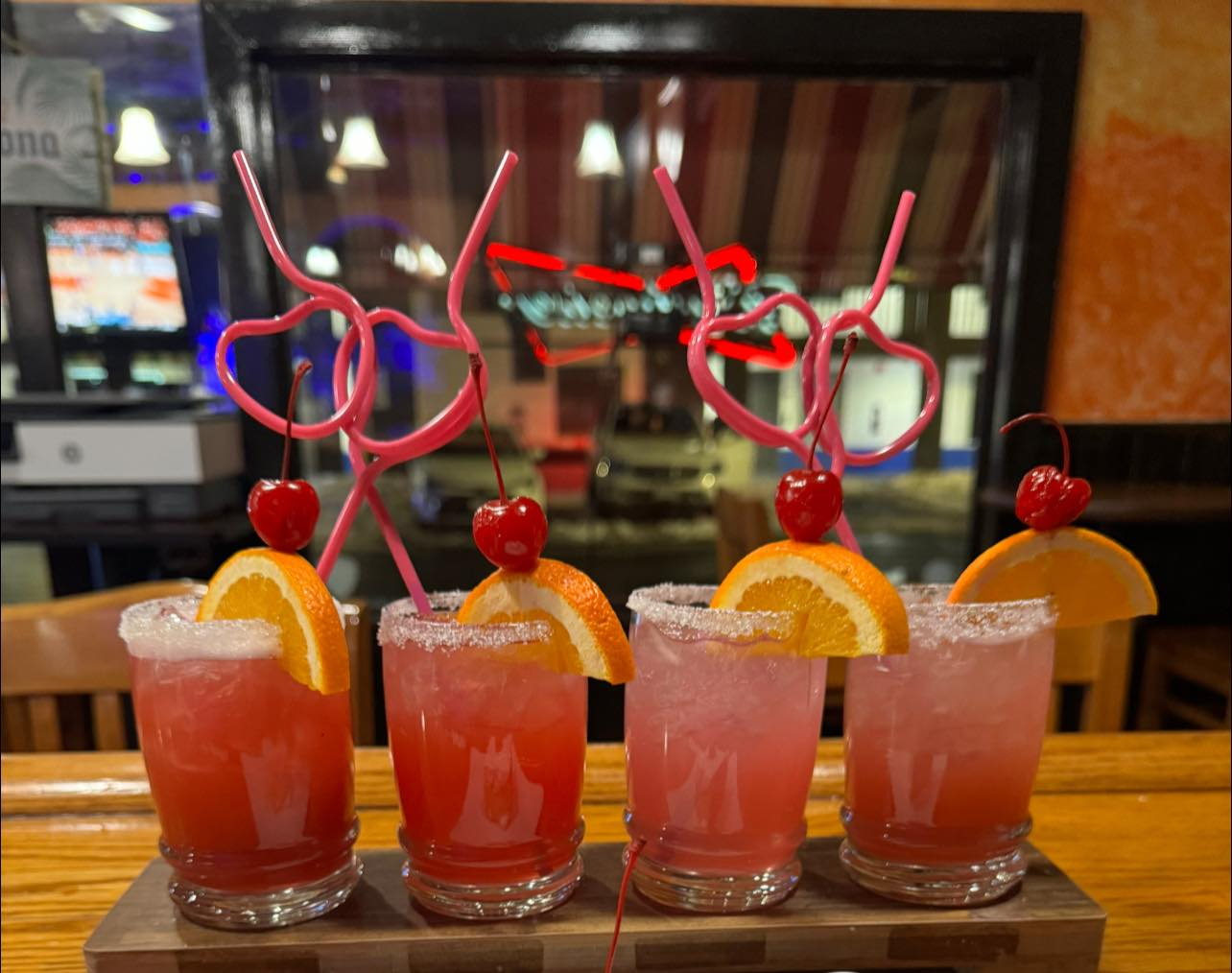 Four pink cocktails with orange slices and cherries on a bar tray, decorated with heart-shaped swizzle sticks.