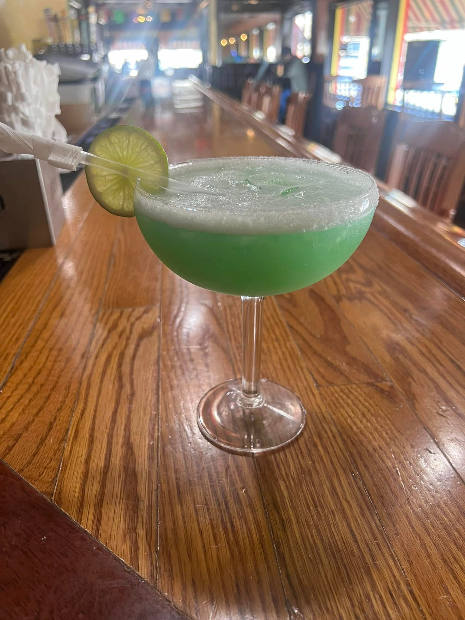 Green cocktail in a stemmed glass with a lime slice, set on a wooden bar counter.