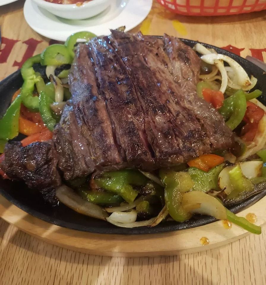 Grilled steak over sautéed peppers and onions on a black plate with rice at the edge of a table