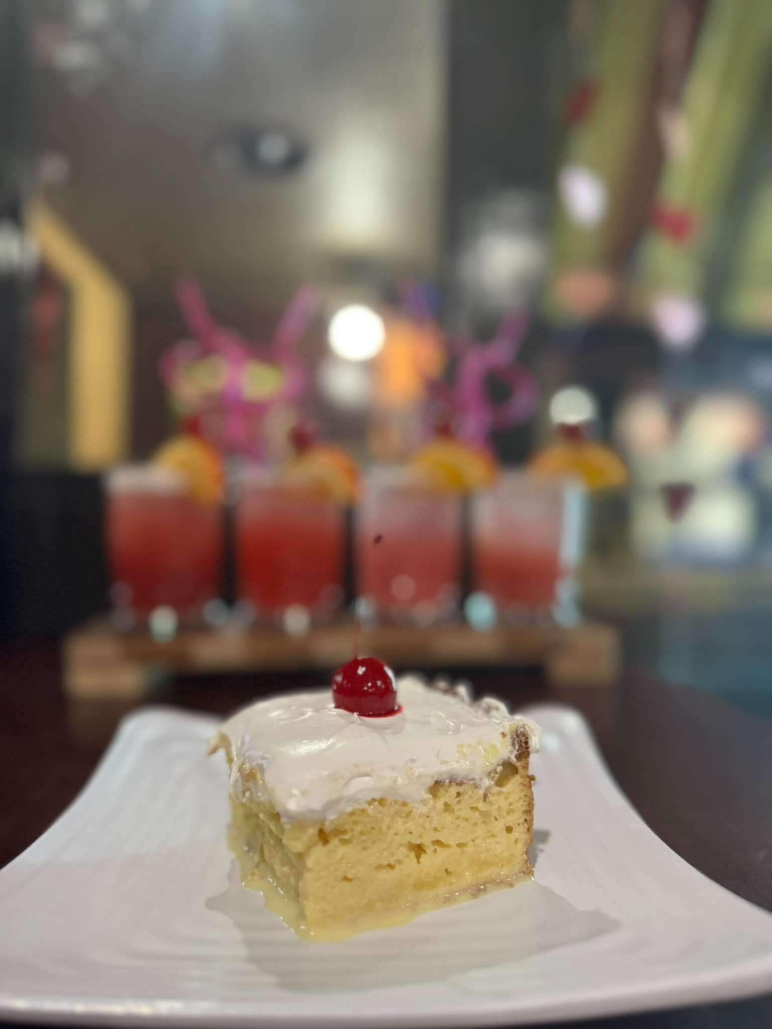 Slice of yellow cake with white frosting and a cherry on a plate, with colorful drinks blurred in the background
