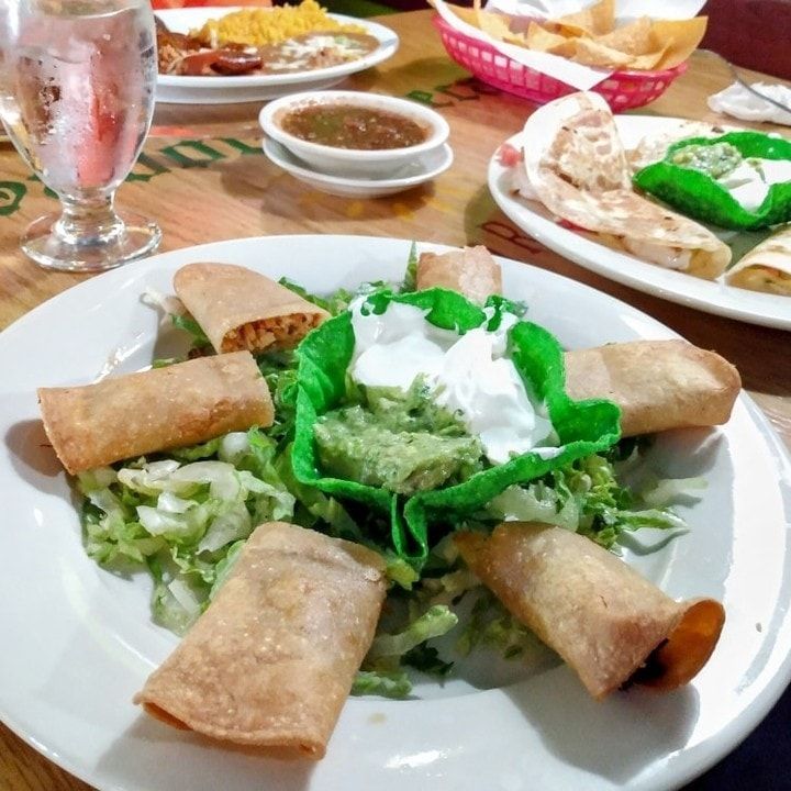 Plate of taquitos on lettuce with guacamole and sour cream, on a dining table with other dishes in the background