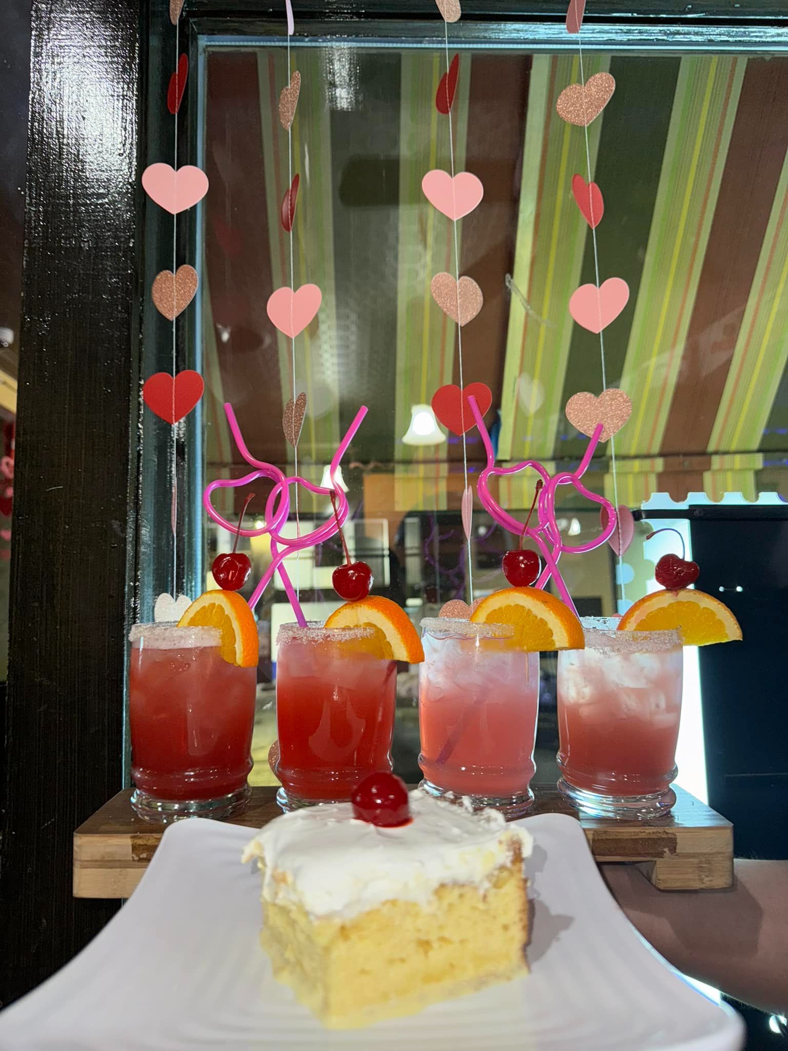 Pink drinks with heart straws and a cake slice on a table by a window.