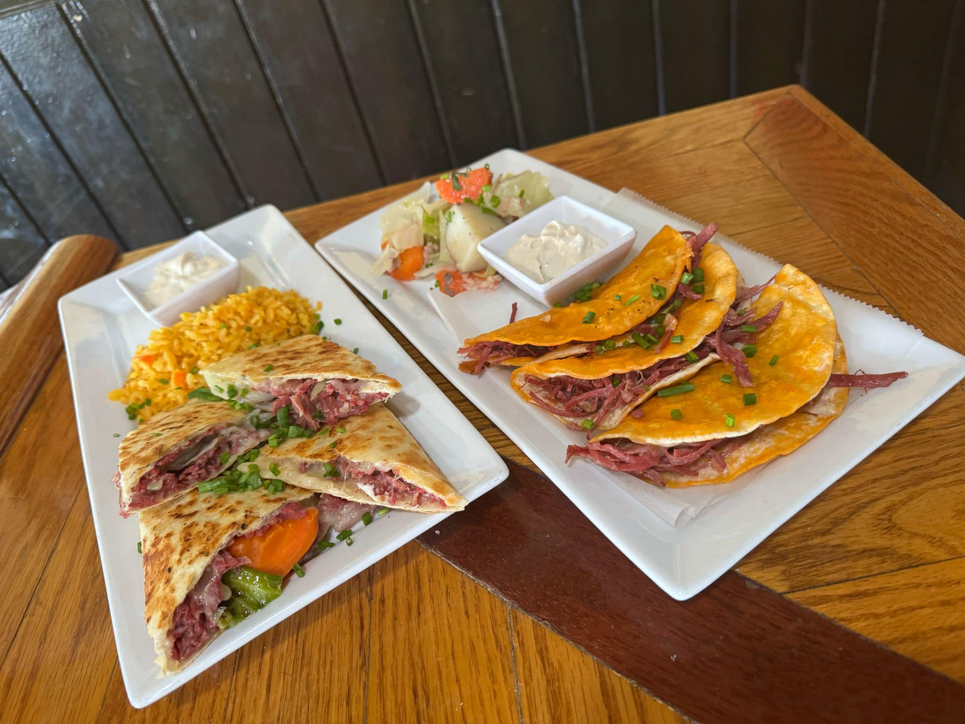Two plates of quesadillas and rice on a wooden table with small cups of salsa and sour cream.