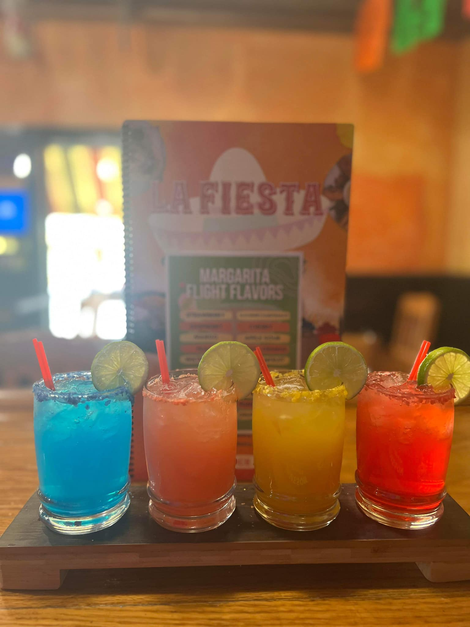 Four colorful cocktails with lime slices on a tray in a dim bar setting.