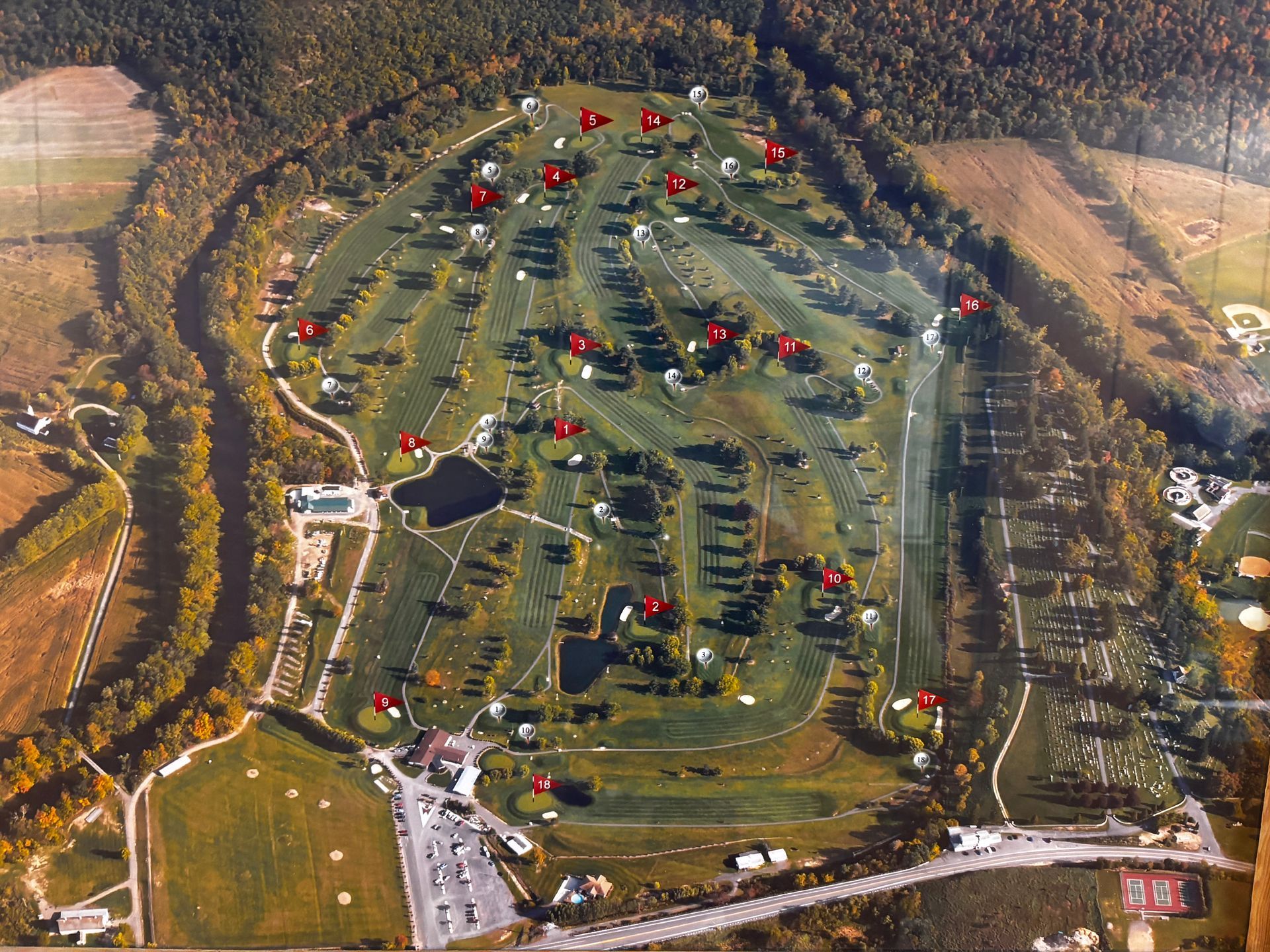 Aerial view of a golf course with red pin flags marking hole locations. The course is surrounded by trees and fields.