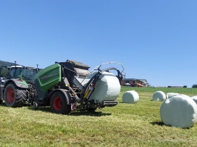 Traktor mit angebauter Ballenwickler wickelt einen Heuballen auf einem Feld unter blauem Himmel.