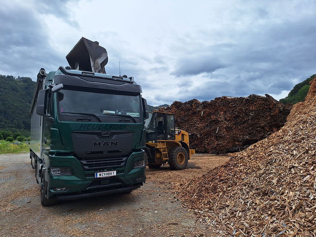 Grüner LKW wird im Freien von einem Lader mit Holzspänen beladen.