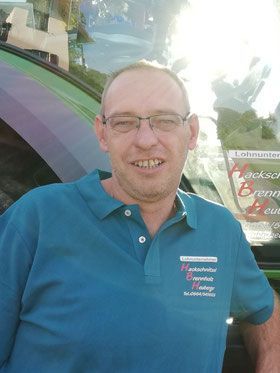 Ein Mann in blauem Poloshirt und Brille lächelt neben einem Fahrzeug mit Firmenlogo.