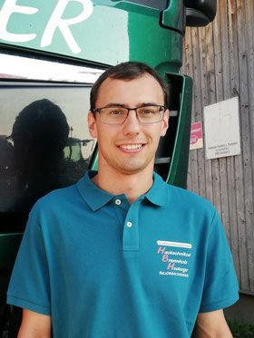 Ein Mann in einem türkisfarbenen Poloshirt lächelt vor einem grünen Lastwagen.
