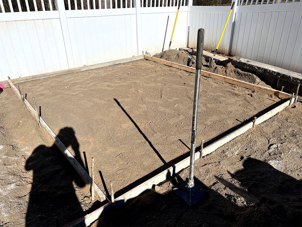 Freshly leveled dirt foundation within white fenced yard, with corner stakes and string lines marking the perimeter