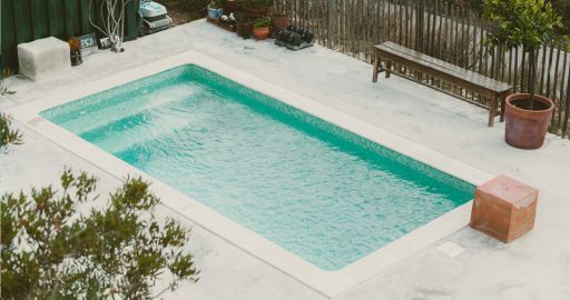 Rectangular turquoise swimming pool in a paved backyard with bench, potted plants, and fence