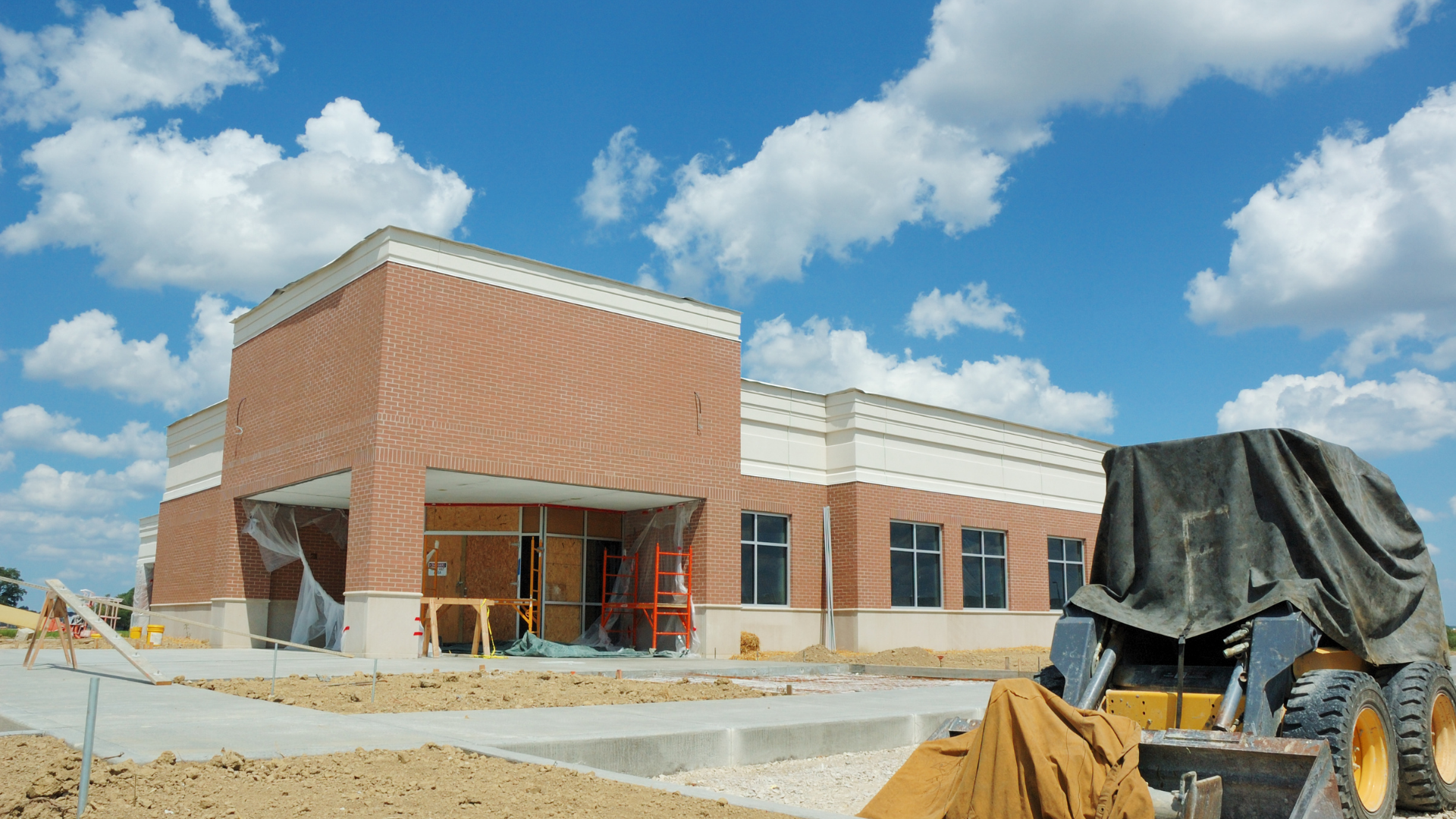 Brick building under construction with scaffolding, blue sky, and a covered vehicle in the foreground