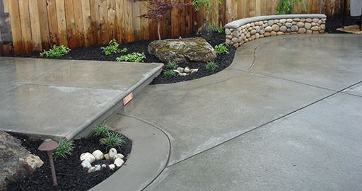 Curved concrete patio and walkway beside a wooden fence, stone wall, and small landscaped planting beds
