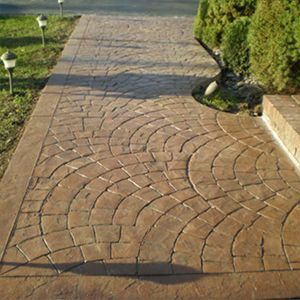 Brick-patterned walkway with curved paving stones beside grass and shrubs in sunlight