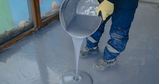 Person pours gray floor leveling compound onto a concrete floor indoors.