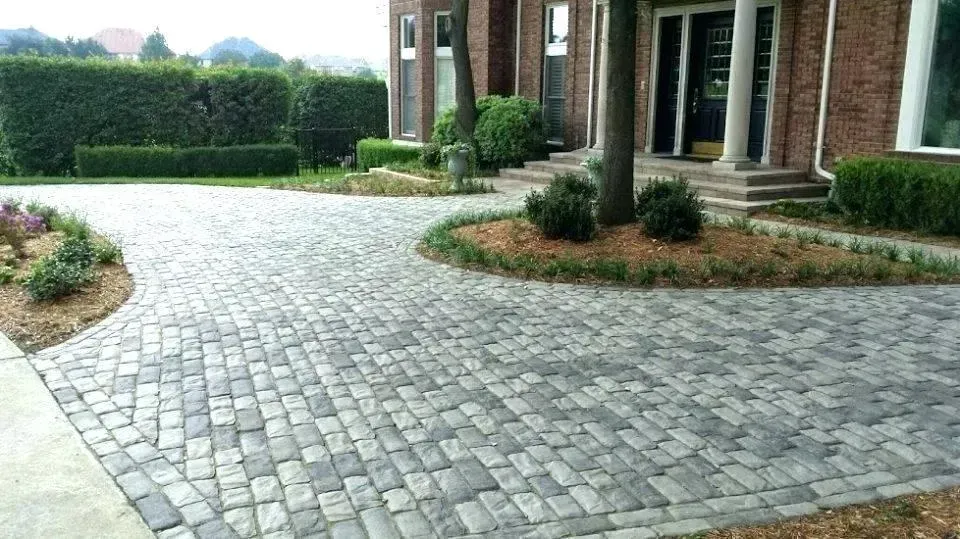 Paved stone driveway beside a brick house with landscaped shrubs and a hedge in the background