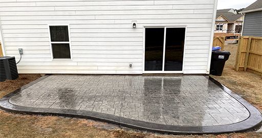 Concrete patio with a curved dark border beside a white house and sliding glass doors