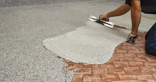 Worker spreading light gray coating over a red brick driveway with a trowel