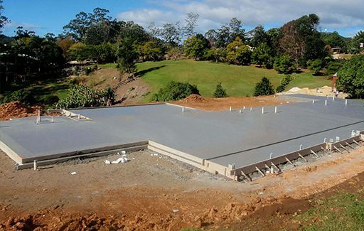 Concrete foundation slab under construction on a grassy site with trees and dirt surrounding it