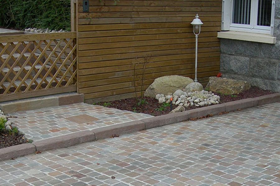 Paved driveway beside a wooden house with a small landscaped border and garden lamp