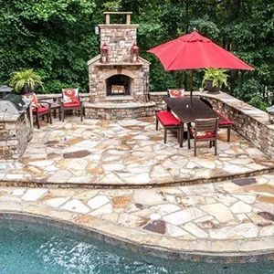 Stone patio with outdoor brick fireplace, red umbrella, chairs, and a pool edge surrounded by greenery
