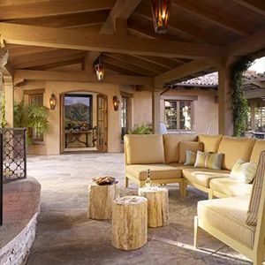 Covered patio with tan sectional seating, log tables, and warm wood beams overlooking a garden courtyard