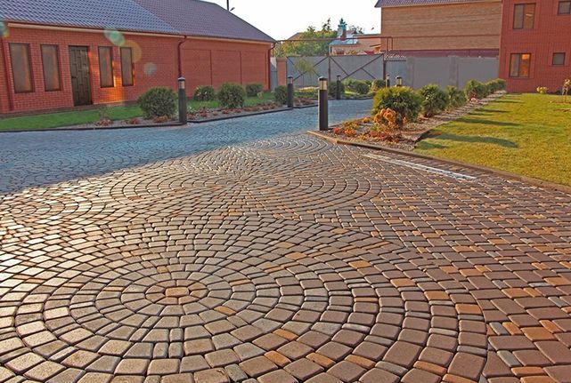 Curved cobblestone courtyard between red brick buildings with landscaped grass and shrubs.