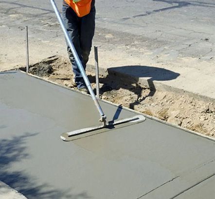Worker smoothing fresh concrete sidewalk with a long-handled screed outdoors
