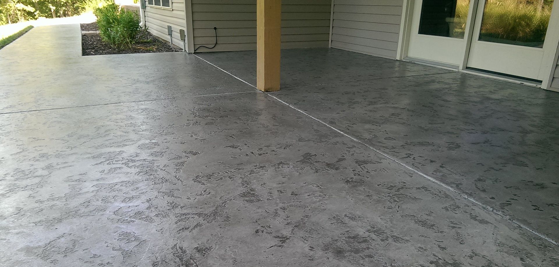 Covered concrete patio with a central support post, garage doors, and a small garden area beyond