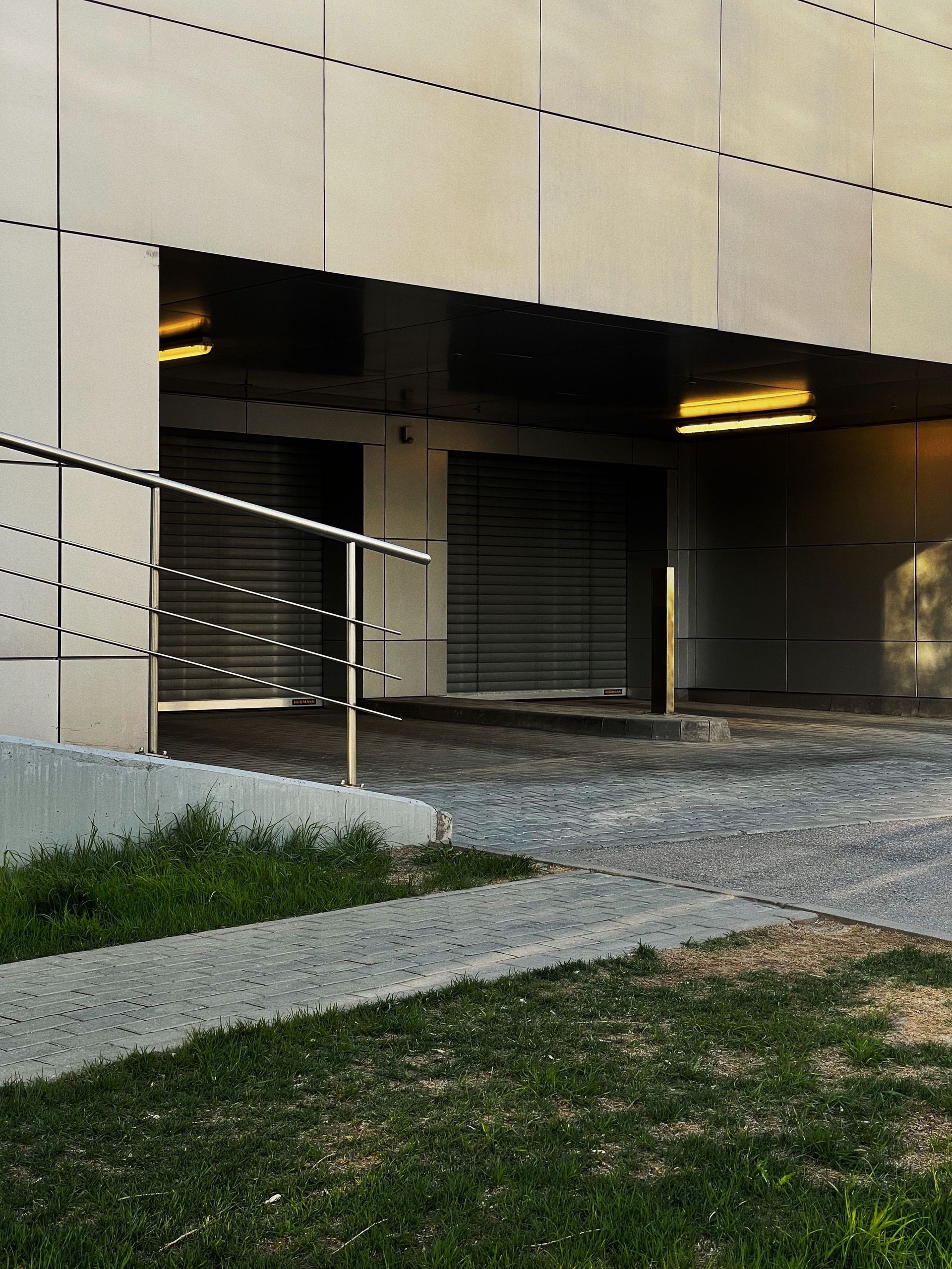 Building entrance with a shuttered doorway, lit overhang, and ramp beside gravel and grass.