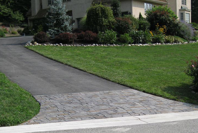 Curved driveway with asphalt and stone pavers leading to a house surrounded by green landscaping