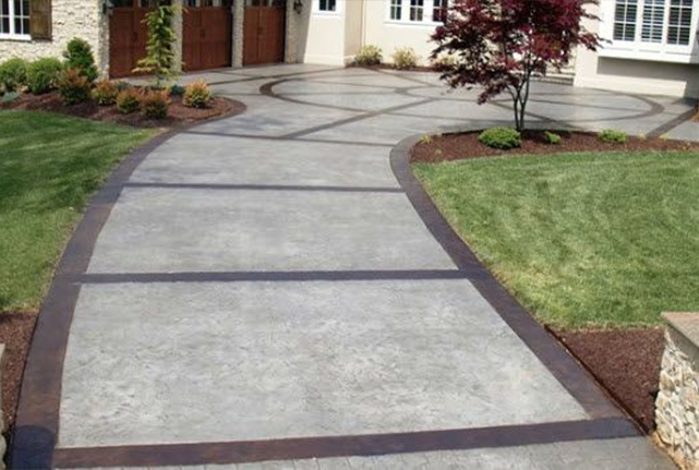 Curved concrete walkway with dark borders leading to a house entrance, bordered by lawn and landscaping.