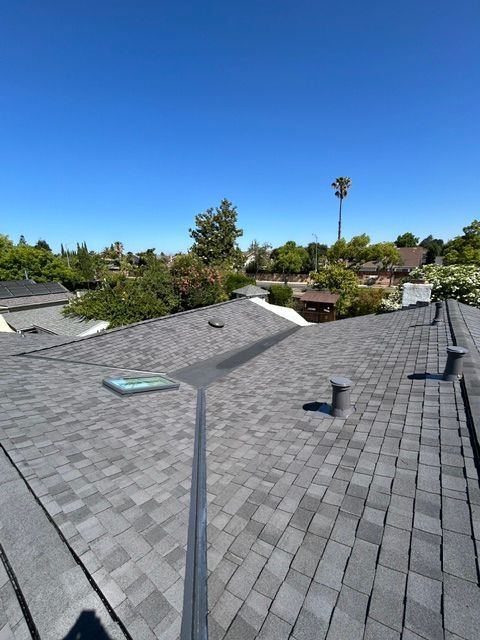View of a gray asphalt shingle roof on a house, clear blue sky, trees, and a cell tower in the distance.