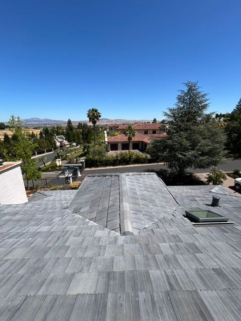 Gray shingle roof with skylight, overlooking suburban homes and distant hills under a blue sky.