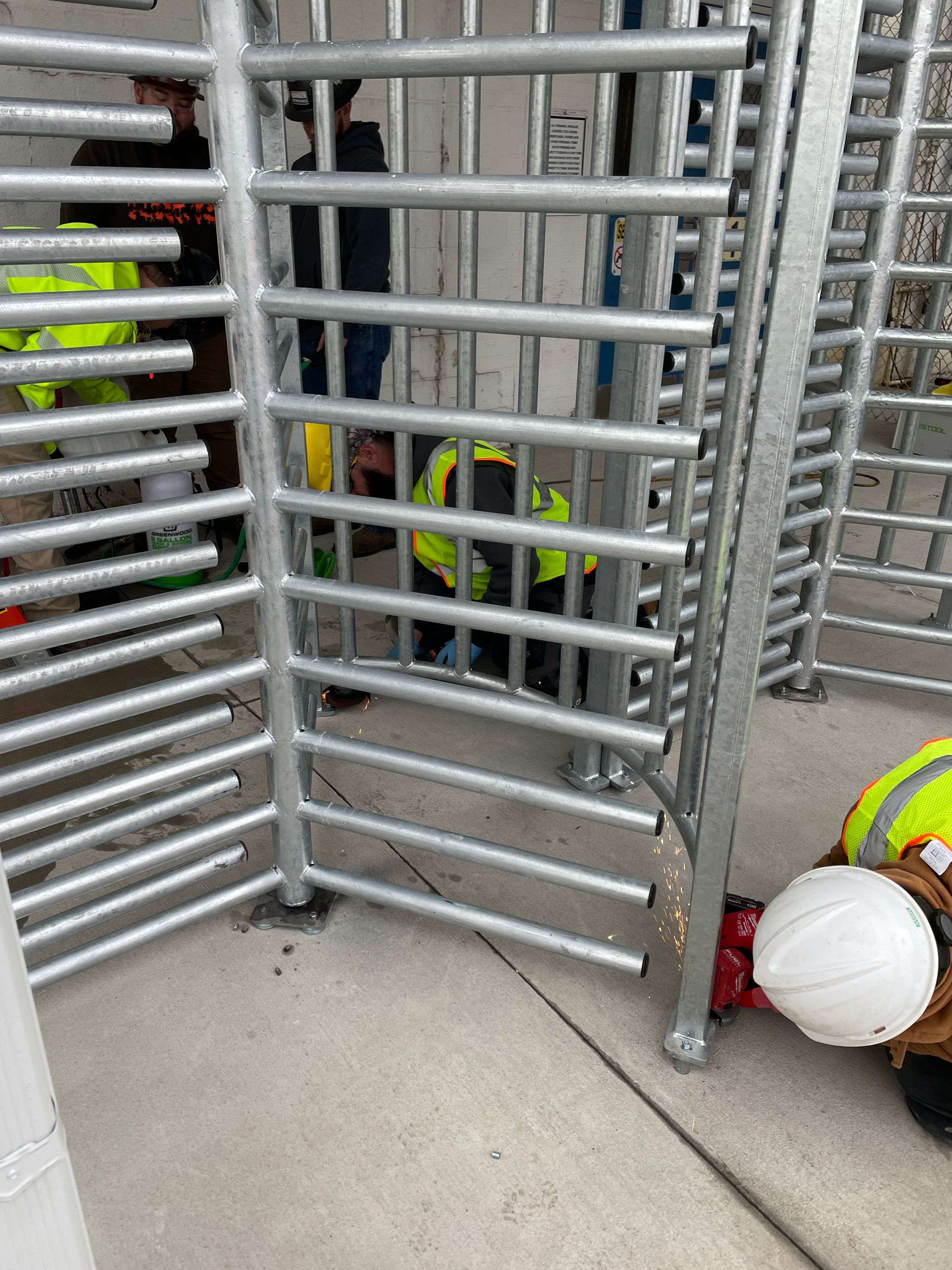 Workers in yellow vests welding metal turnstiles on concrete.